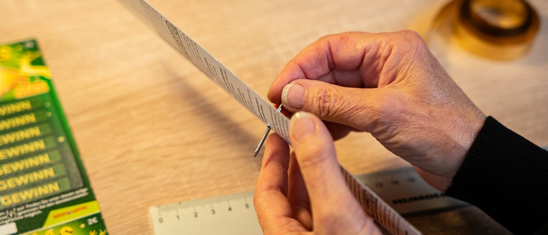 Eine Hand sticht mit einem Nagel durch ein Rubbellos | WestLotto