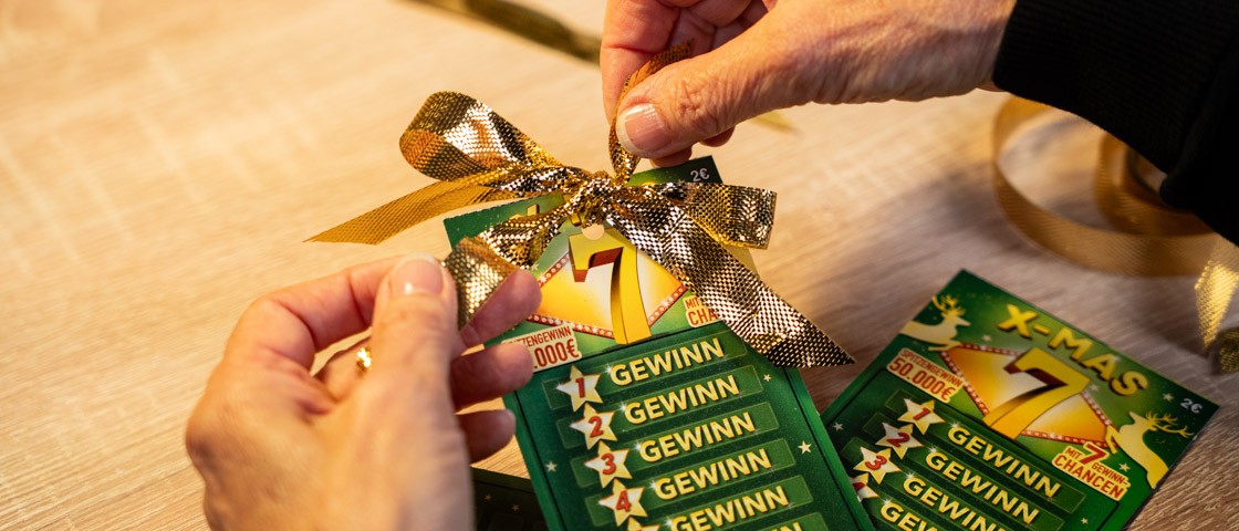 Zwei Hände binden eine goldene Schleife an ein Rubbellos | WestLotto